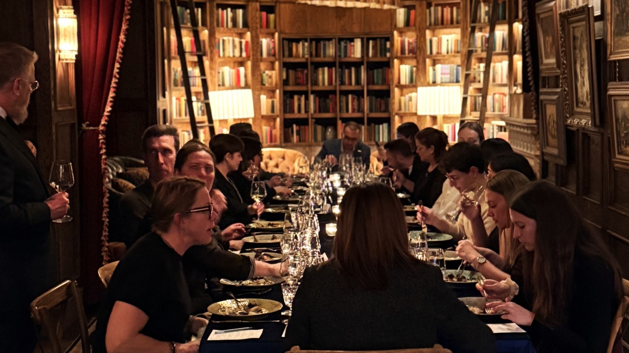 a long dining table set in a library room with books floor to ceiling. table seats are full of guests enjoying food and wine