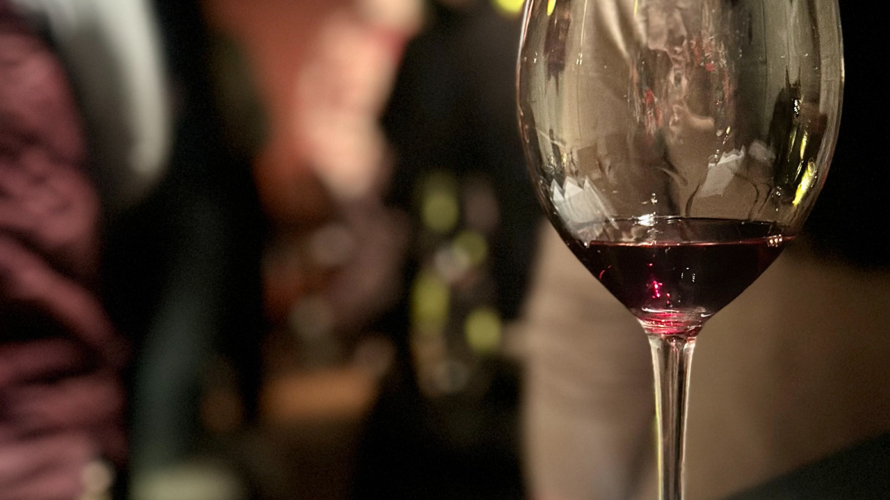 stemmed wine glass with a splash of red wine in a dimly lit room with people standing around in the background out of focus